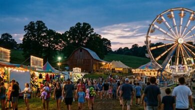 Families walking a county fair midway at dusk with a lit Ferris wheel and a barn in the background