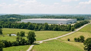 A large windowless industrial building sits behind a treeline buffer above rolling Middle Tennessee pasture and hay fields, with transmission towers along the property edge (illustrative)