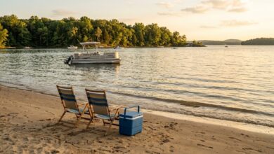 Two empty beach chairs and a cooler on a sandy lakeshore at golden hour, with a pontoon boat offshore and a wooded far bank.